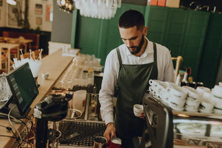 Barista in a green apron steams milk at the counter of a café