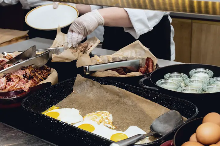 Hand in a disposable glove placing sausages in the chafing dish of the breakfast buffet, bacon and fried eggs in front
