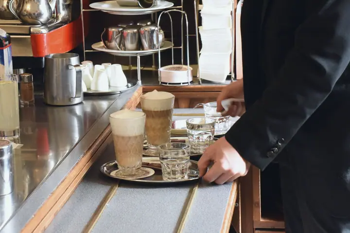 Waiter carries a tray with a Melange and a glass of water in a coffee house