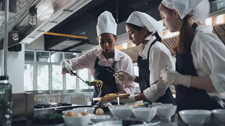 Three apprentices in chef's hats, hair covers and gloves plate up in the training kitchen