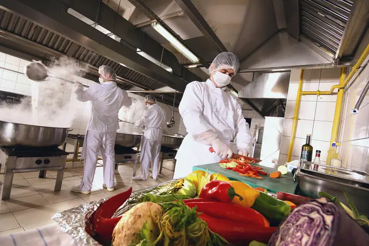 Cooks with hair nets, face masks and gloves in a large kitchen; at the front, vegetables are cut on a green board