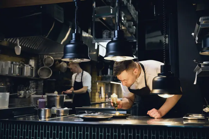 Chef plating up under heat lamps in a dark professional kitchen