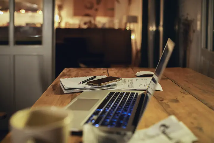 Wooden table in a restaurant after closing: an open laptop, a notepad with a pen, a phone and printed lists, behind it the warm light of the empty dining room