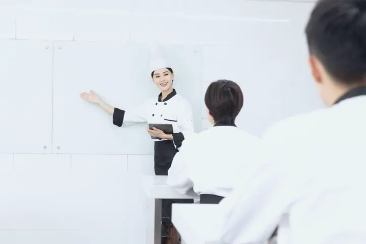 A teacher in a chef's hat explains something at the board in a cookery school